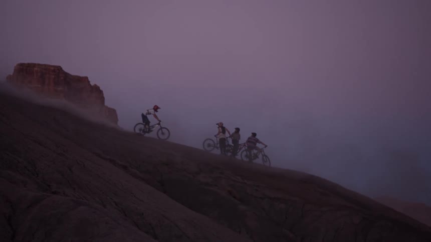 An extreme mountain biker performs a wheelie trick on a steep, dusty desert ridge while his friends watch at dusk