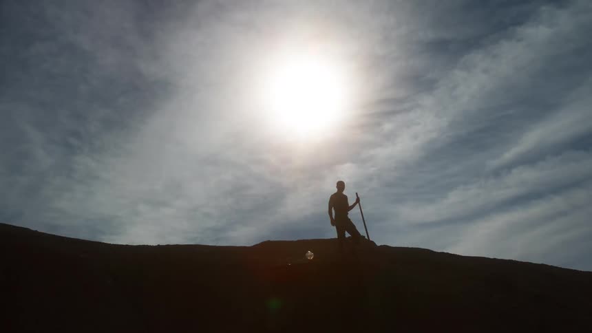 A silhouetted man holding a shovel slowly walks down a large hill against the bright sun and cloudy sky