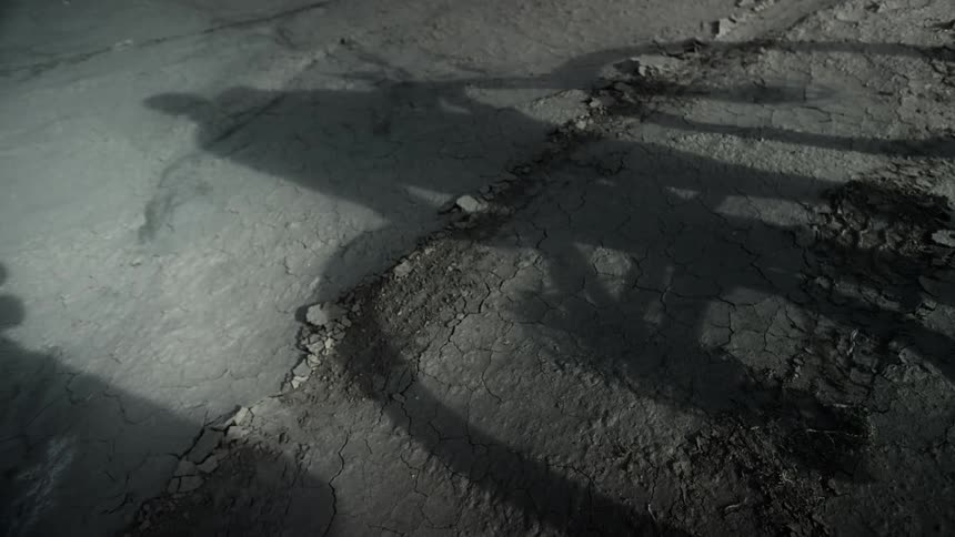 The shadow of a cyclist begins to pedal their bike across a dry, cracked, muddy ground