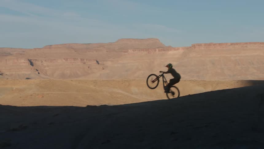 A mountain biker does a long wheelie in slow motion on a desert ridge with canyons in the background