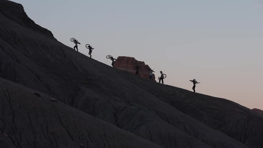 A group of silhouetted mountain bikers carry their bikes up a steep, barren hill at dusk