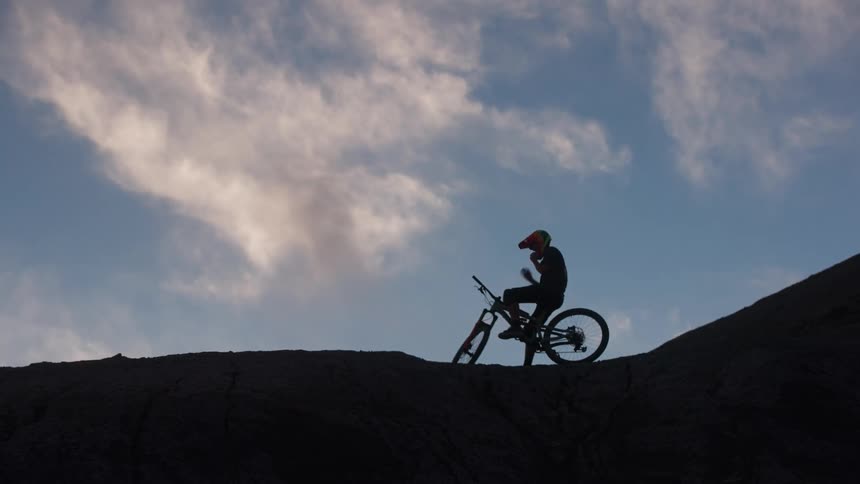 Silhouette of a mountain biker sitting on a bike and putting on a helmet on a rocky ridge at dawn