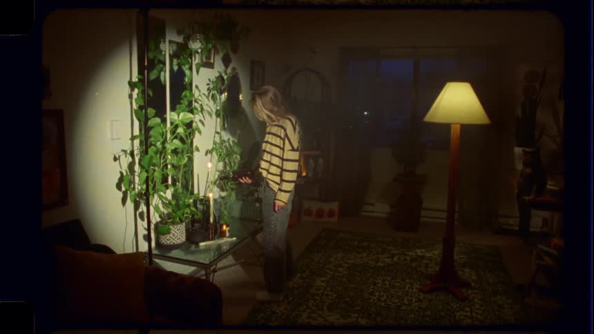 Vintage film-style shot of a young woman standing in a spotlight in her cozy, plant-filled living room at night