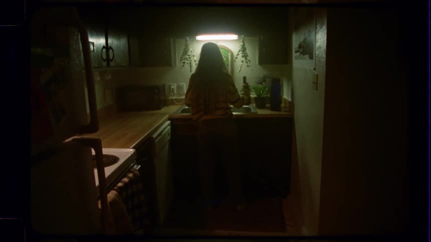A woman stands at the sink in a dimly lit kitchen in this vintage-style footage