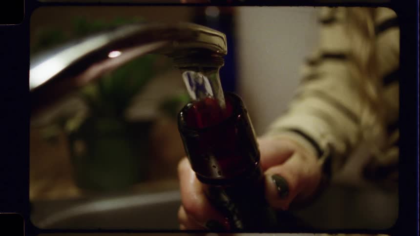A vintage-style shot shows a person filling a dark glass bottle with water from a tap