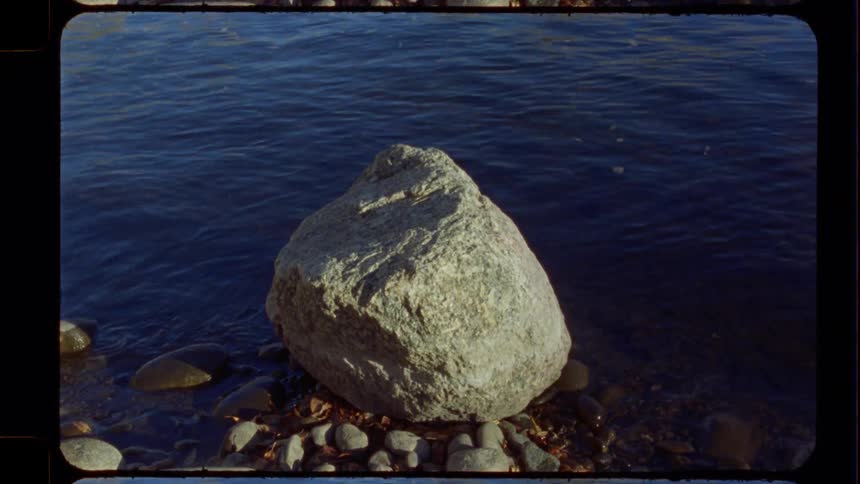 A large, sun-drenched boulder rests on a pebbly shore beside gently rippling dark blue water in a vintage film shot