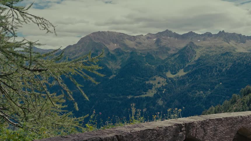 A serene view of a vast mountain range and lush green valley from behind a stone wall on a cloudy day