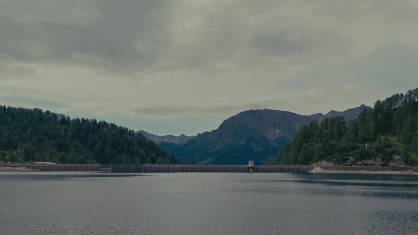 A wide scenic shot of a large dam and reservoir nestled between forested mountains under a cloudy sky
