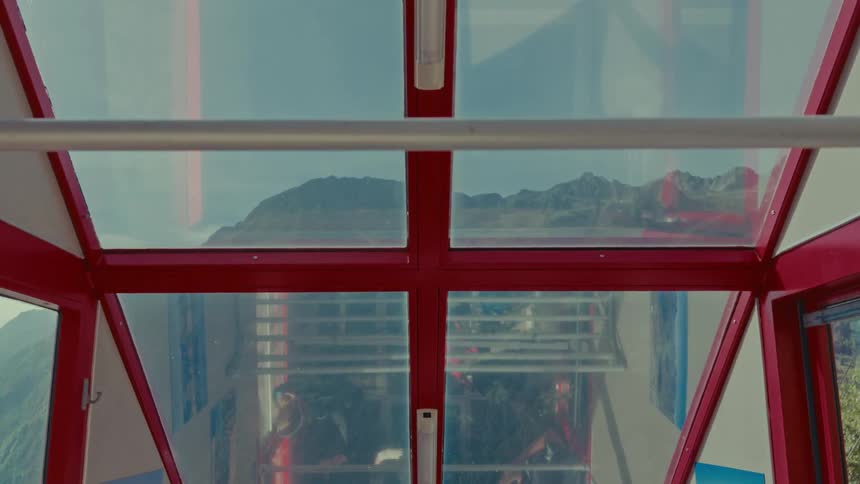Looking up through the glass roof of a red cable car at the mountains as another car passes by