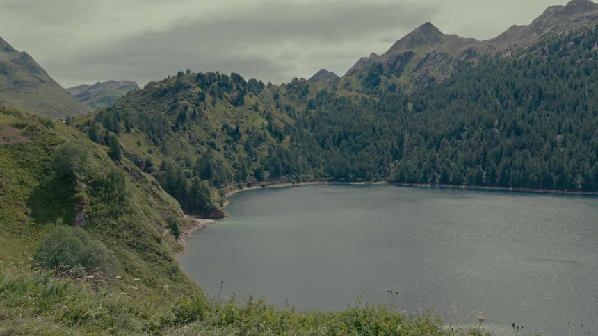 A scenic, wide shot of a calm, tranquil mountain lake surrounded by lush green hills and forests on a cloudy day