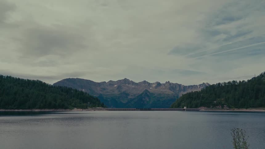 A serene and expansive view of a mountain reservoir and dam nestled between forested hills under a dramatic, cloudy sky