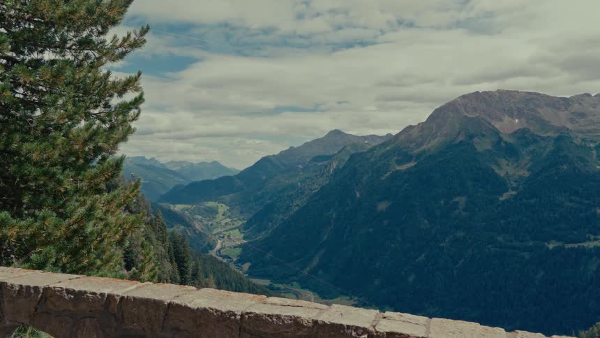 A stunning, picturesque view of a vast mountain valley with green forests unfolds from a stone lookout on a cloudy day