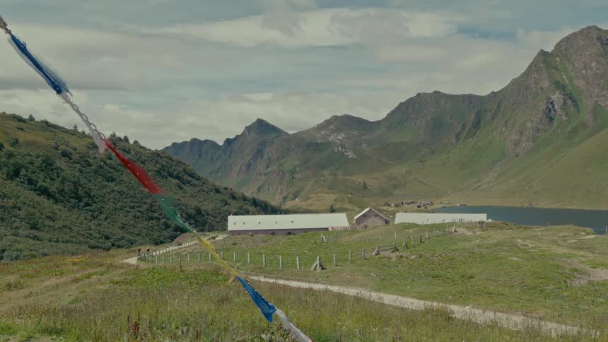 Tibetan prayer flags wave gently in the wind overlooking a serene mountain valley with a path leading to a lakeside farm