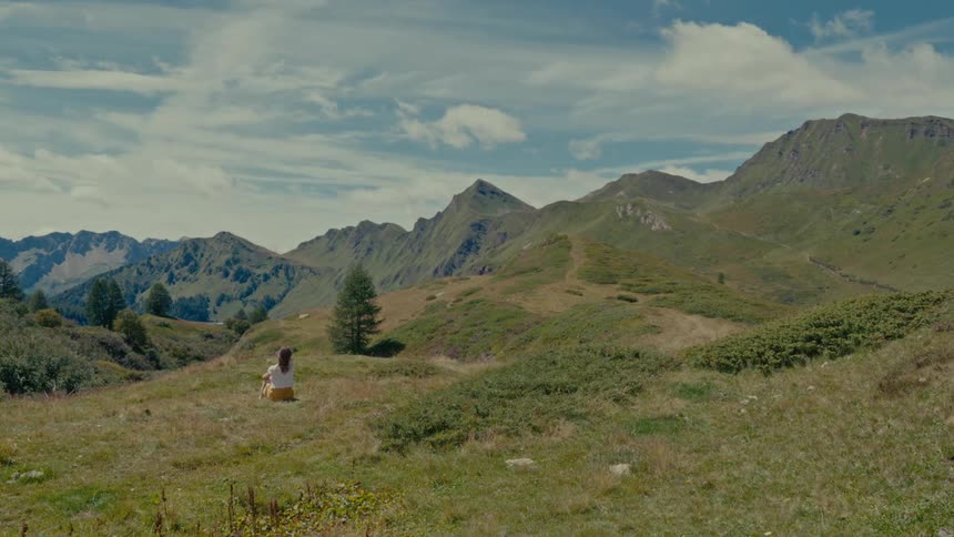 A solitary woman sits in a vast green mountain valley, peacefully contemplating the serene natural scenery
