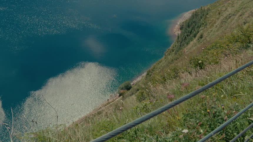 A high-angle view from a steep, grassy cliffside looks down onto a sparkling, deep blue lake on a sunny day
