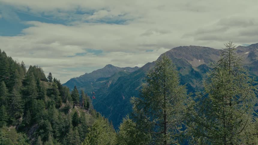 A red cable car ascends a lush green mountainside against a backdrop of majestic peaks and a cloudy sky