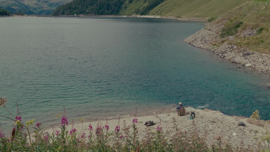 A lone angler sits peacefully on the rocky shore of a tranquil mountain lake, framed by wildflowers