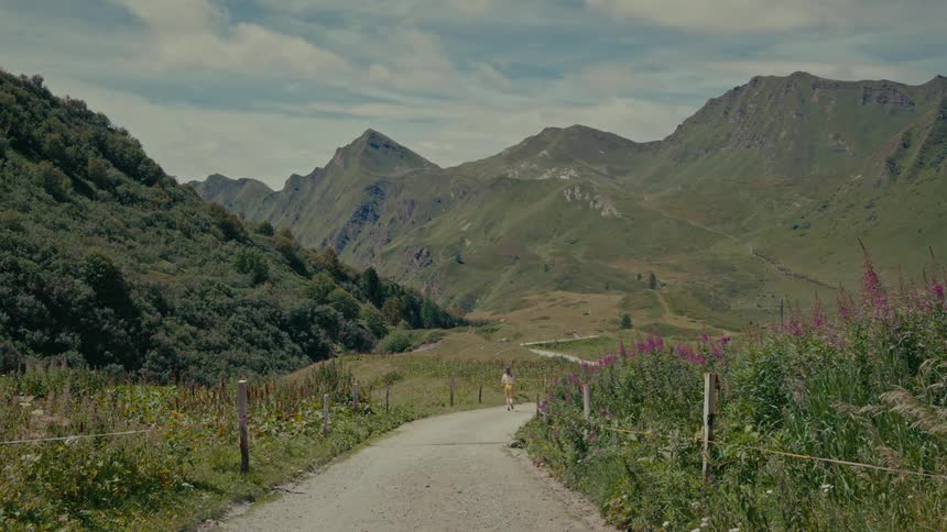 A young woman walks down a winding dirt road through a vibrant green mountain valley on a sunny day