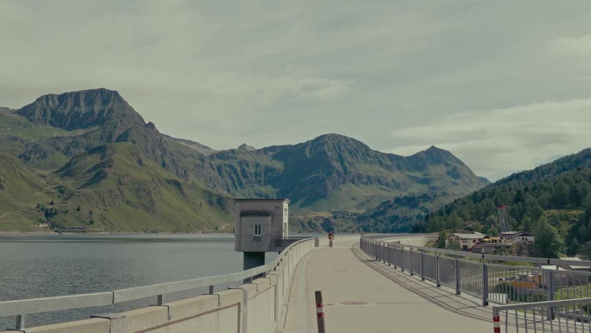 A lone cyclist rides across a massive dam high in the mountains next to a serene lake