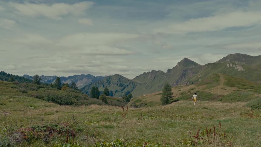 A woman in a yellow skirt walks through a picturesque mountain valley under a cloudy sky