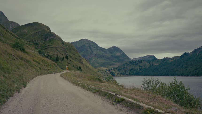 A solitary woman takes a scenic walk along a lakeside path surrounded by vast green mountains under a cloudy sky