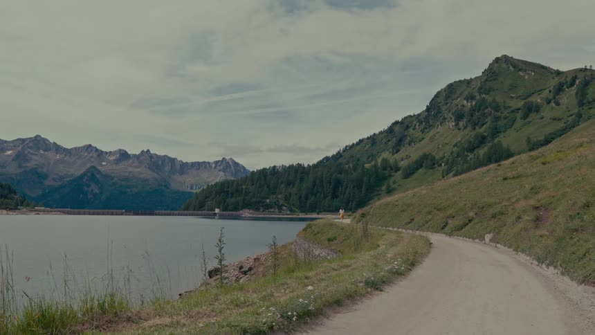 A woman and her dog enjoy a scenic walk on a dirt road alongside a vast mountain lake with a dam in the background