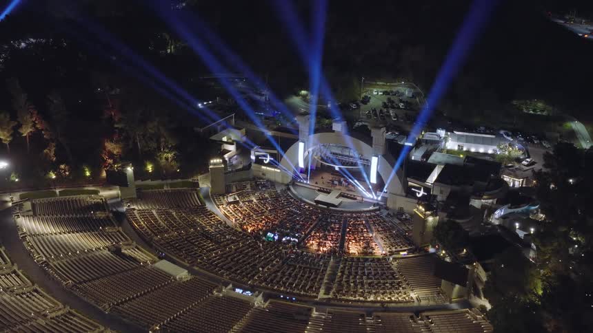 An aerial view captures a live concert at the iconic Hollywood Bowl at night, with blue spotlights illuminating the sky
