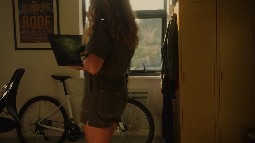 A woman standing in her sunlit apartment works on her laptop next to her bicycle before walking off-screen