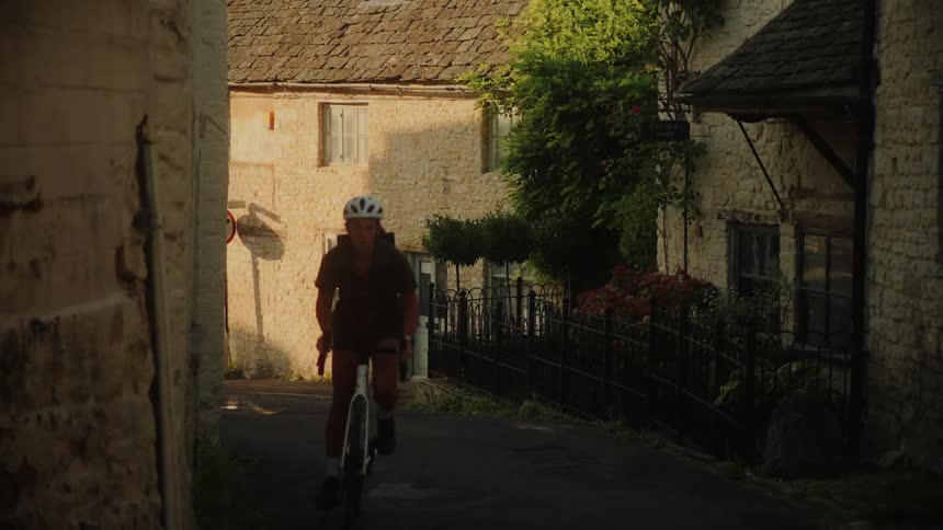 A woman wearing a helmet rides her bicycle up a narrow street through a quaint stone village during golden hour
