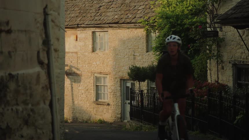 A female cyclist rides her bike in slow motion up a narrow street past quaint stone houses in a European village