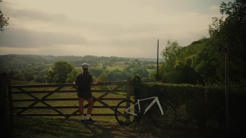 A cyclist pauses their ride to lean against a wooden gate and soak in the beautiful view of the rolling green countryside