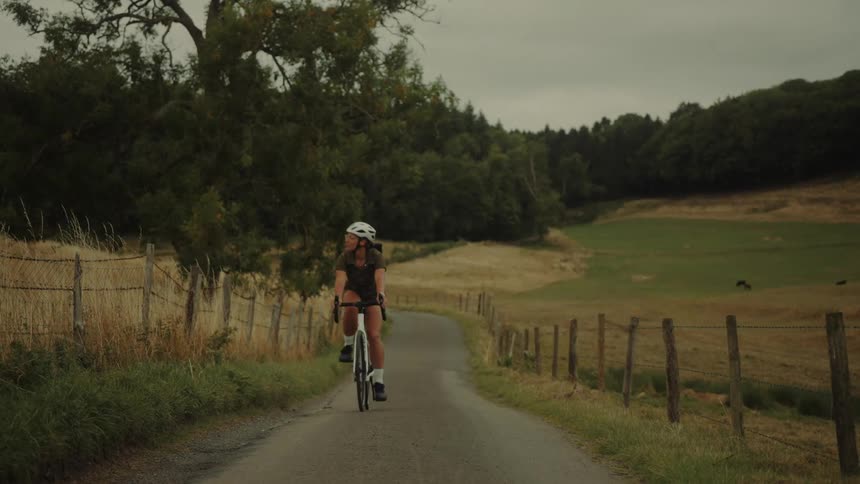 A female cyclist wearing a helmet pedals towards the camera on a quiet, winding country road surrounded by green fields and trees
