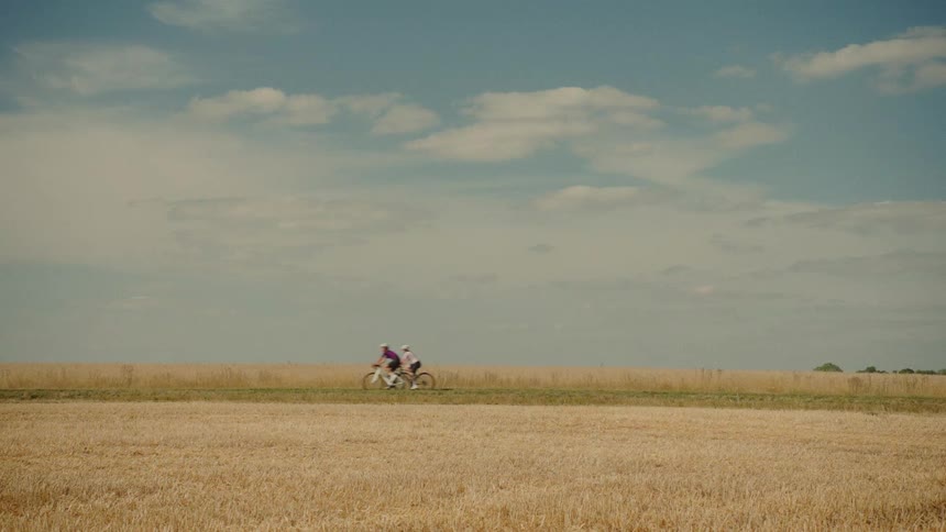 A pair of cyclists enjoy a peaceful ride through the countryside past a vast golden wheat field under a blue summer sky