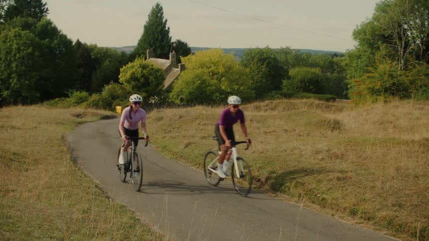 Two female cyclists ride up a winding country road through rolling hills on a sunny day