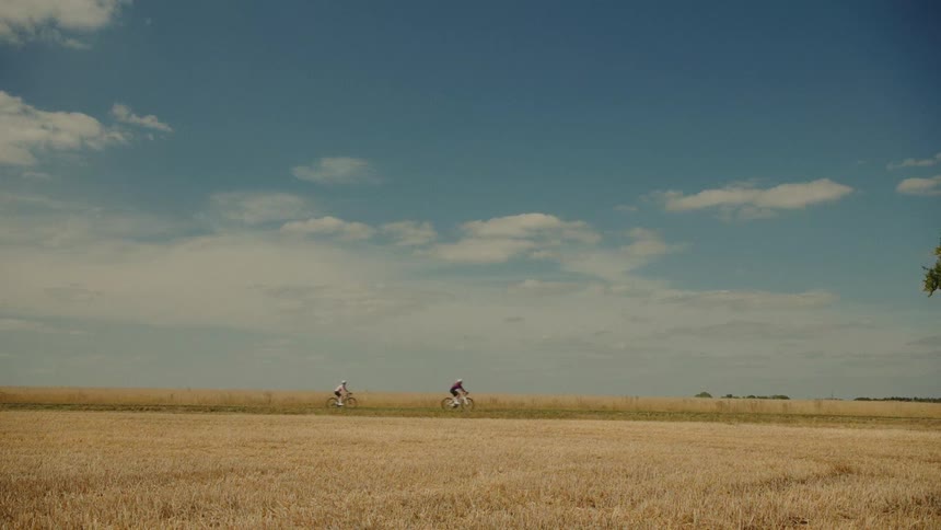 Two cyclists ride along a path through a golden harvested field under a vast blue sky on a sunny day
