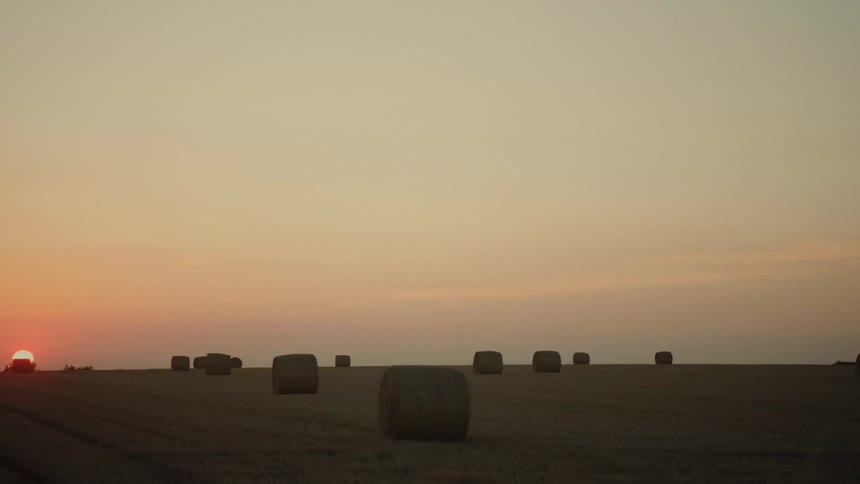 A beautiful, warm sunset glows over a vast rural field dotted with large hay bales