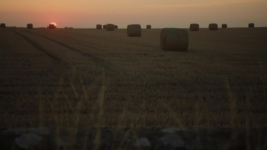 A serene wide shot shows large hay bales scattered across a vast field as the sun sets on the horizon