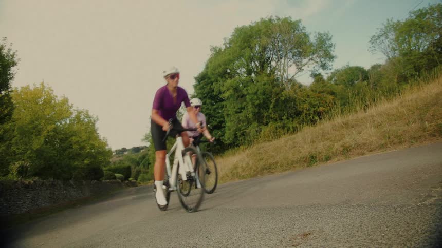 Two female cyclists pedal hard as they climb a steep hill on a rural country road