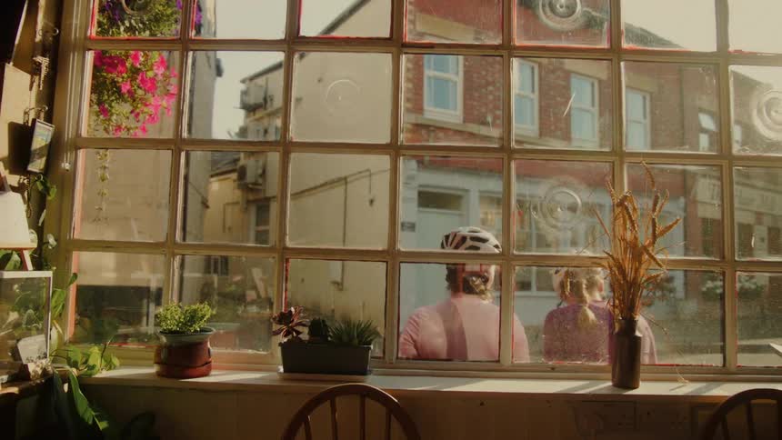 A view from a sunlit cafe window shows two cyclists in helmets stopping on a quaint town street