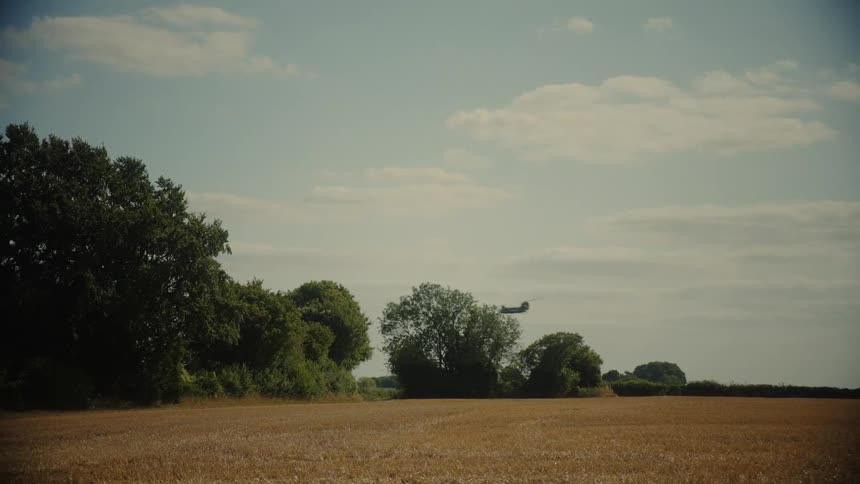 A military Chinook helicopter flies low over a golden field and treeline on a sunny day