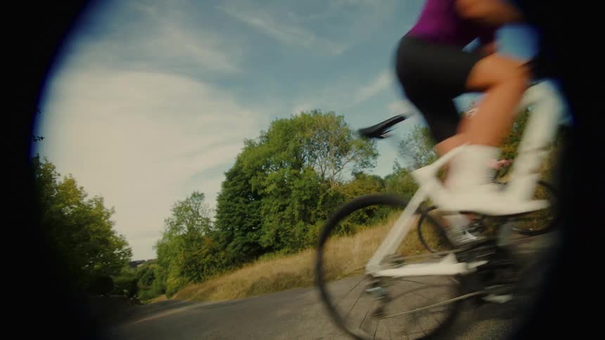 A low-angle, vignetted shot captures two female cyclists pedaling up a steep country road on a sunny day