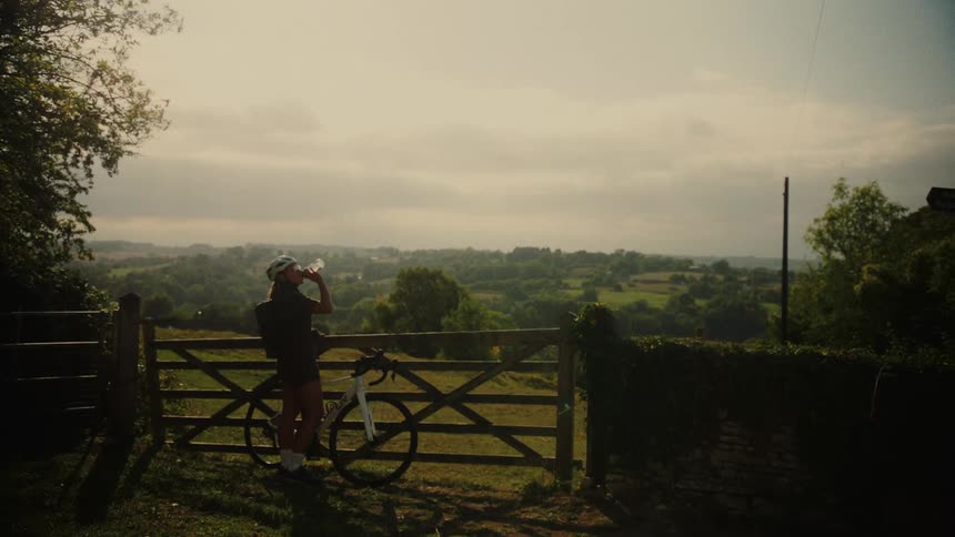 A female cyclist takes a refreshing water break by a wooden gate, admiring the beautiful, rolling green countryside view
