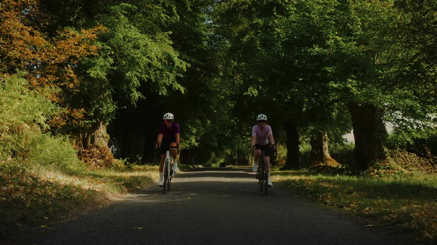 Two female cyclists ride their bikes towards the camera down a beautiful tree-lined country lane