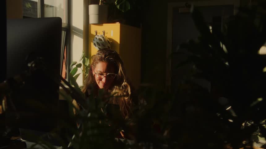 A focused woman with long hair and glasses works at her computer in a warm, sunlit office