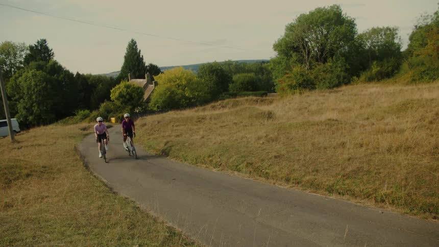 Two female cyclists ride up a winding country road through rolling hills on a sunny day