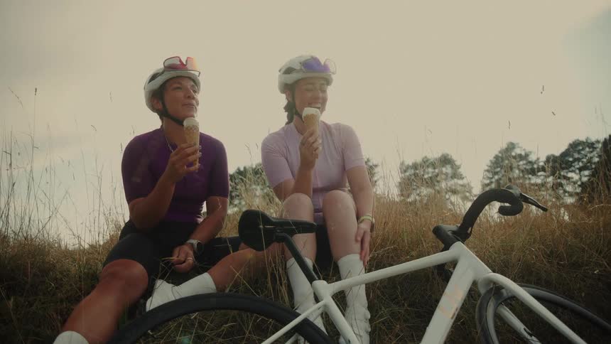 Two female cyclists laugh together while eating ice cream cones and taking a break in a grassy field