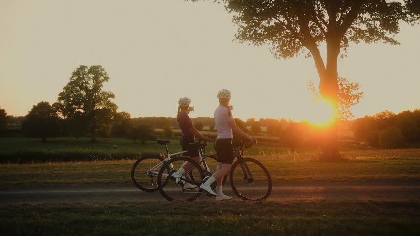 Two female cyclists take a break from their ride to walk their bikes and enjoy the beautiful golden sunset in the countryside
