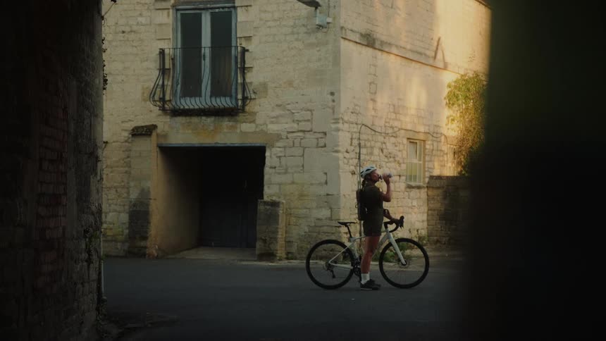 A woman on a bicycle stops for a water break on a quiet street in a rustic European village