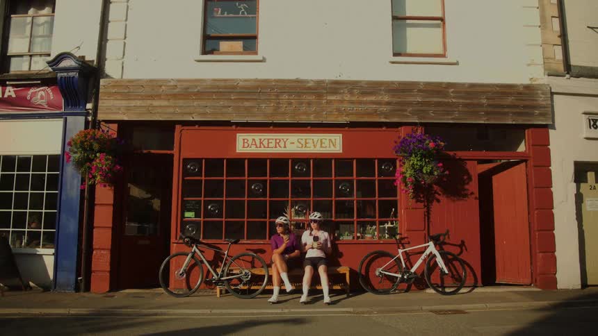 Two female cyclists take a break on a bench, eating and drinking outside a quaint red bakery with their bikes nearby