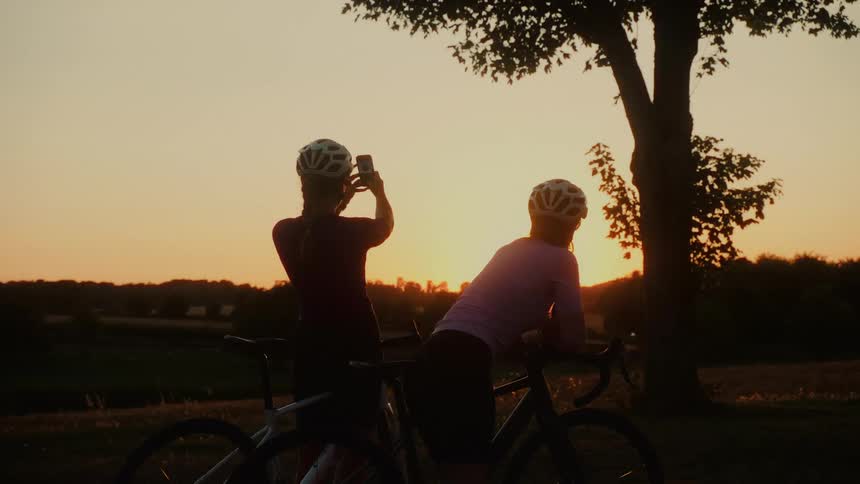 Silhouetted against a golden sunset, two female cyclists pause their ride to capture the scenic countryside view on a phone